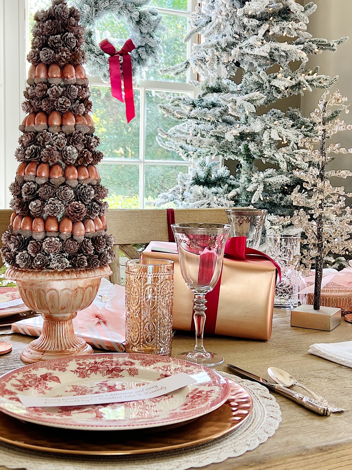 A Christmas table set in cranberry and rose gold colors with a DIY tree, cranberry and white plates, amber and clear glasses, rose gold chargers, and wrapped packages.