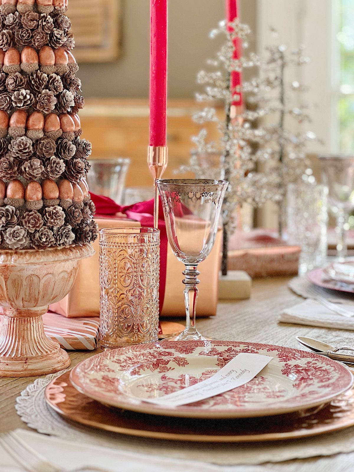 A Christmas table set in cranberry and rose gold colors with a DIY tree, cranberry and white plates, amber and clear glasses, rose gold chargers, and wrapped packages.