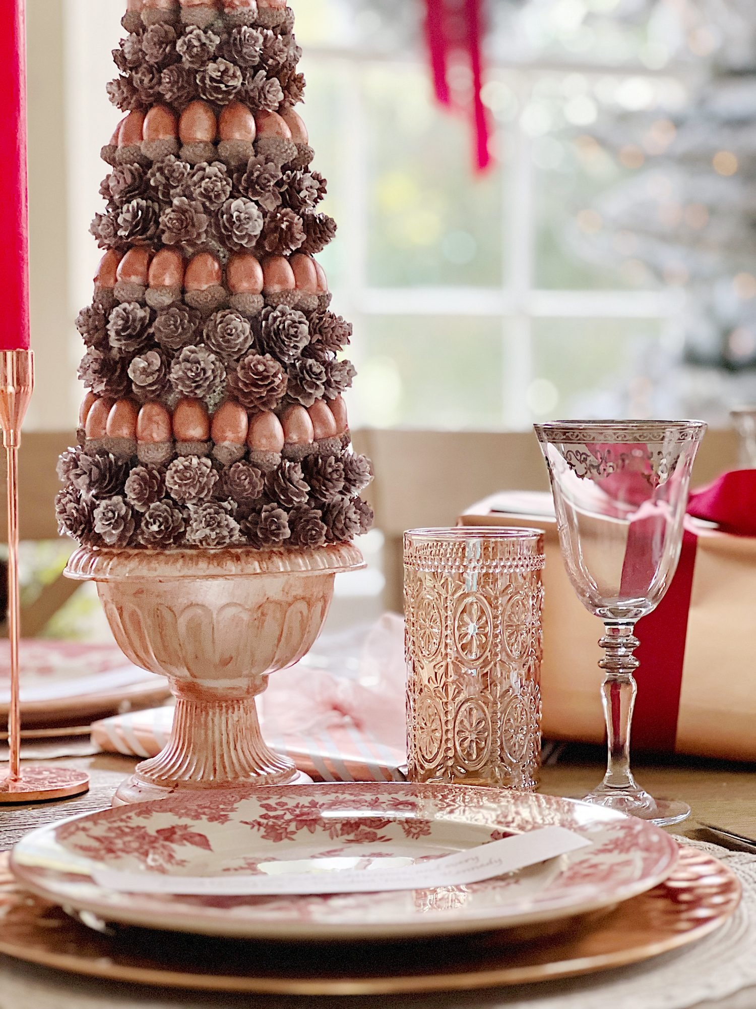 A Christmas table set in cranberry and rose gold colors with a DIY tree, cranberry and white plates, amber and clear glasses, rose gold chargers, and wrapped packages.