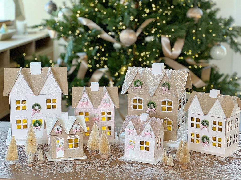 A festive display of gingerbread houses is arranged on a table with mini trees and nutcracker figurines, all set against a decorated Christmas tree background.