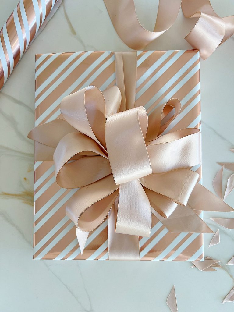 Instructions to tie a bow with ribbon on your Christmas gifts using rose gold metallic wrapping paper and assorted ribbons.