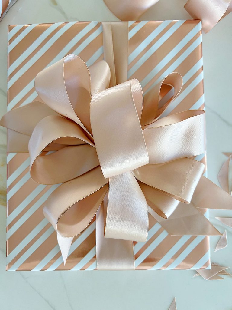 Instructions to tie a bow with ribbon on your Christmas gifts using rose gold metallic wrapping paper and assorted ribbons.