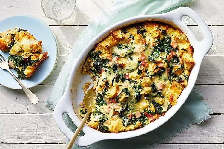 A festive casserole dish with baked strata featuring bread, greens, and melted cheese, perfect for a Nutcracker Christmas. A portion is served on a plate beside the dish, ready to delight your holiday guests.