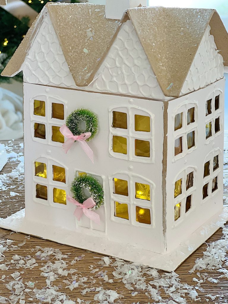 Gingerbread houses made from cardboard and decorated with paint, puff pens, pearls, glitter, tiny wreaths, and pink bows, in a color palette of pink and white.