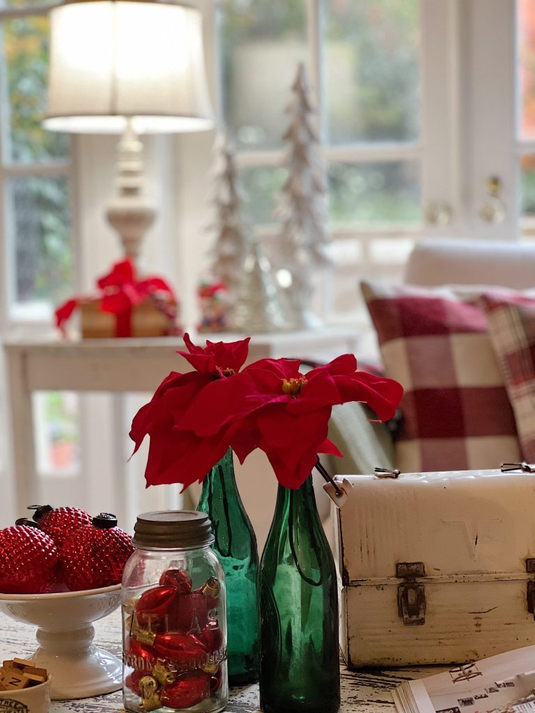A room decorated with red and green holiday accents including poinsettias, a plaid pillow, gifts, green bottles, and ornaments.