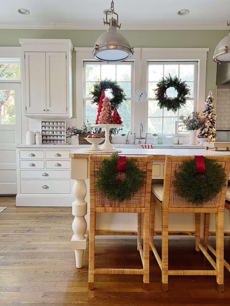 Our white kitchen decorated with wreaths in the windows and on the back of the counter stools, red, green, and white trees, red and white towels, and more.