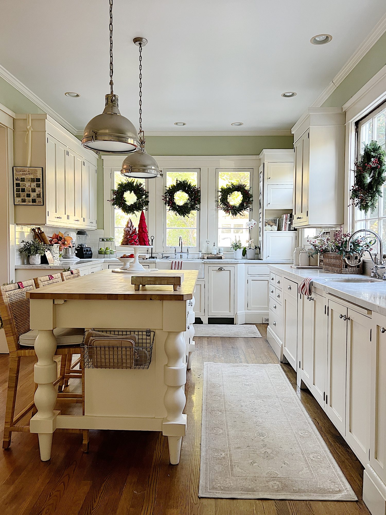Our white kitchen decorated with wreaths in the windows and on the back of the counter stools, red, green, and white trees, red and white towels, and more.