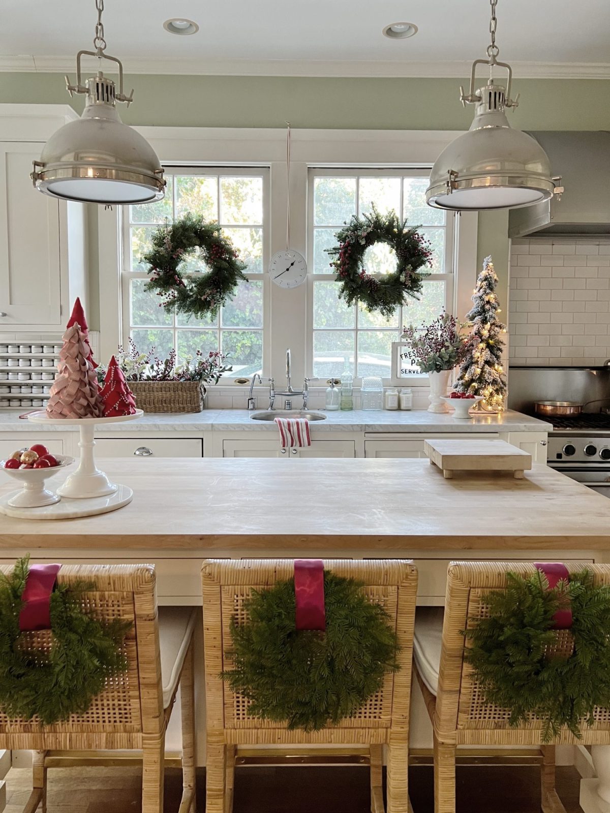 Our white kitchen decorated with wreaths in the windows and on the back of the counter stools, red, green, and white trees, red and white towels, and more.