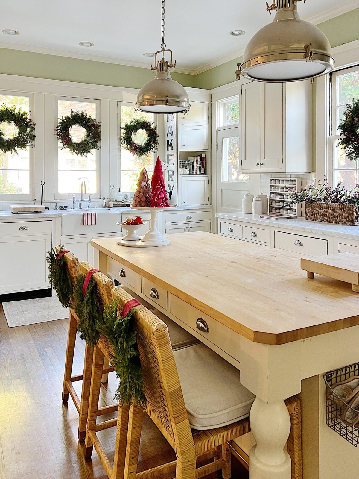 Our white kitchen decorated with wreaths in the windows and on the back of the counter stools, red, green, and white trees, red and white towels, and more.