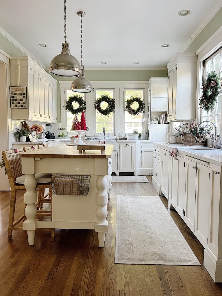 Our white kitchen decorated with wreaths in the windows and on the back of the counter stools, red, green, and white trees, red and white towels, and more.