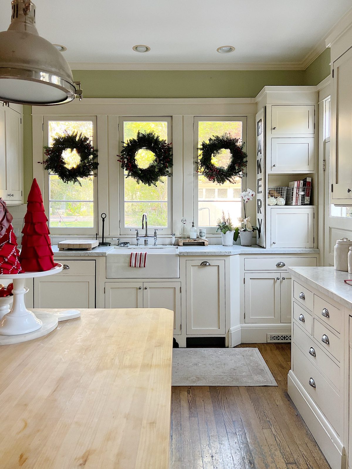 Our white kitchen decorated with wreaths in the windows and on the back of the counter stools, red, green, and white trees, red and white towels, and more.