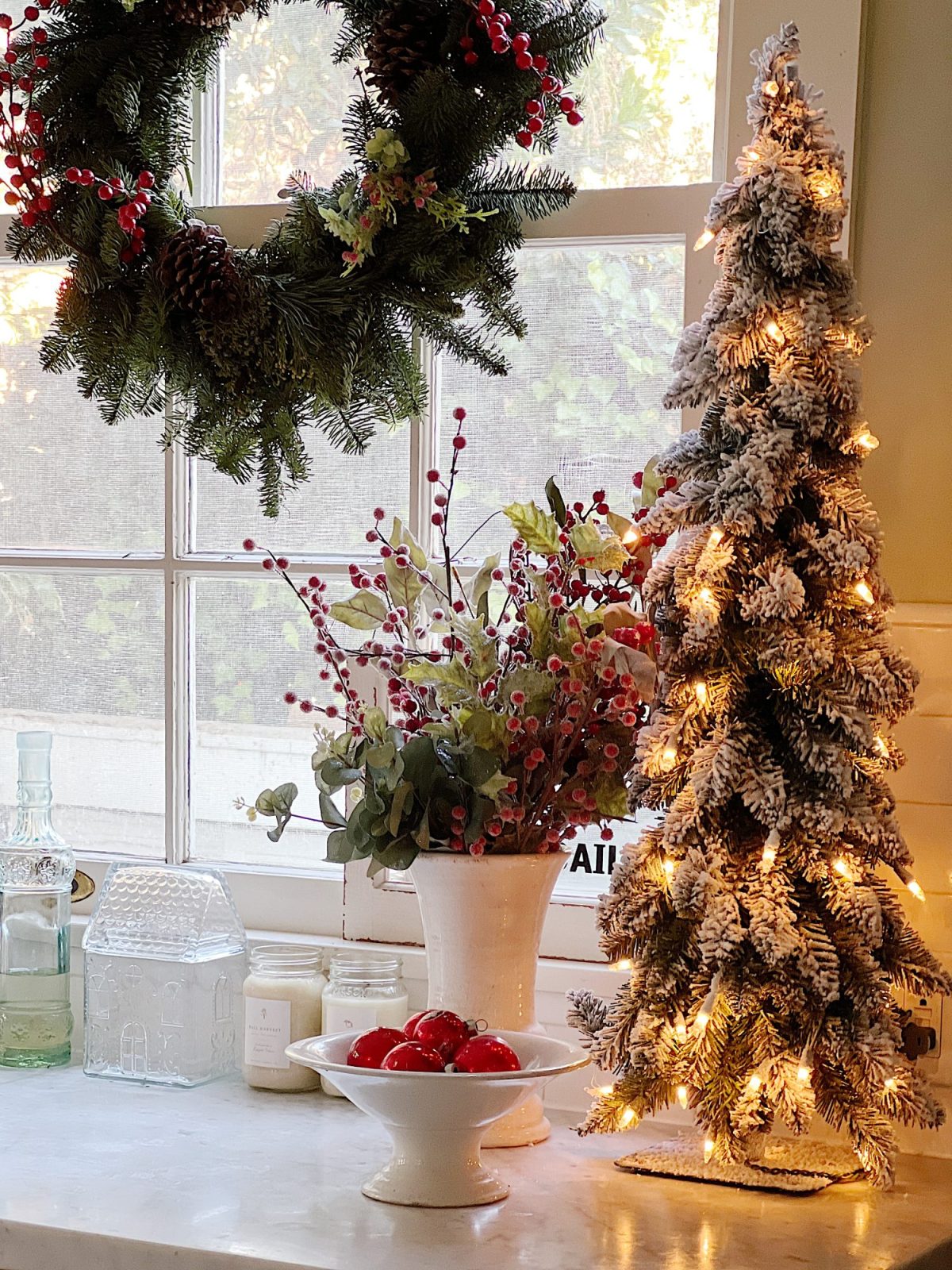 Our white kitchen decorated with wreaths in the windows and on the back of the counter stools, red, green, and white trees, red and white towels, and more.