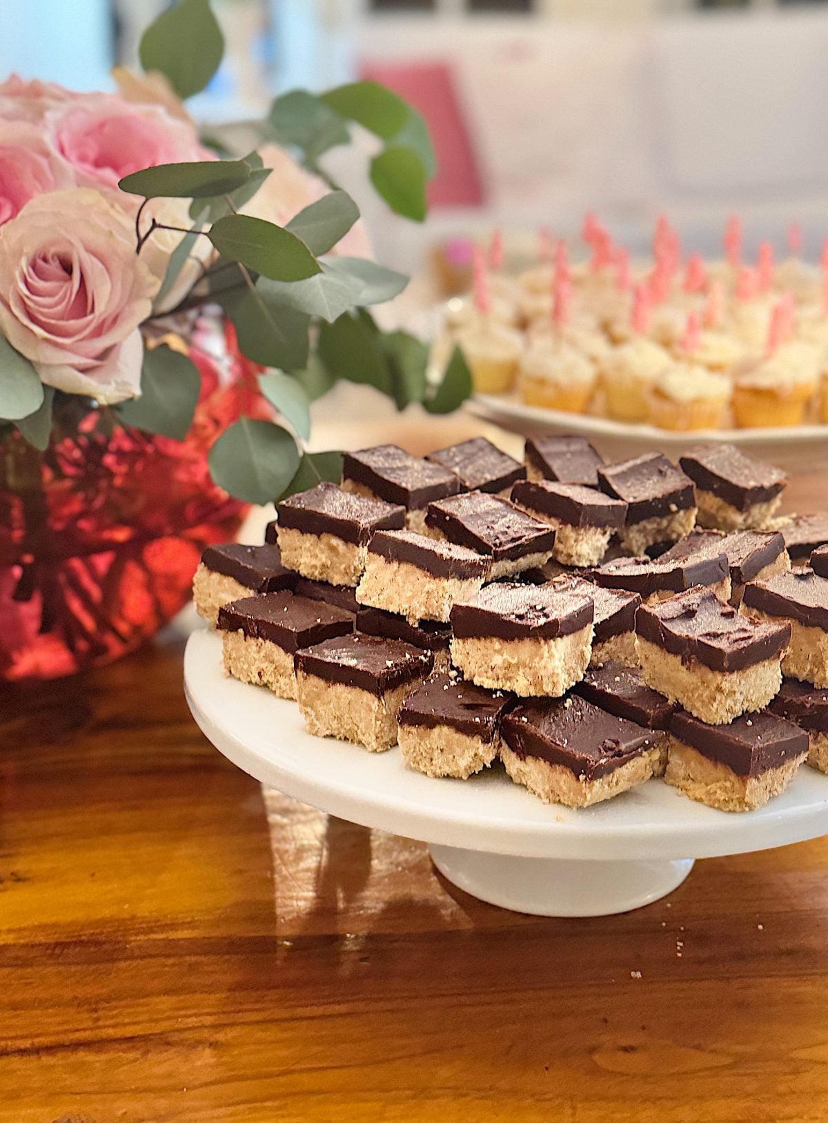 A platter of chocolate-covered square desserts, reminiscent of a decadent peanut butter fudge recipe, sits elegantly on a wooden table with a bouquet of pink roses in the background.