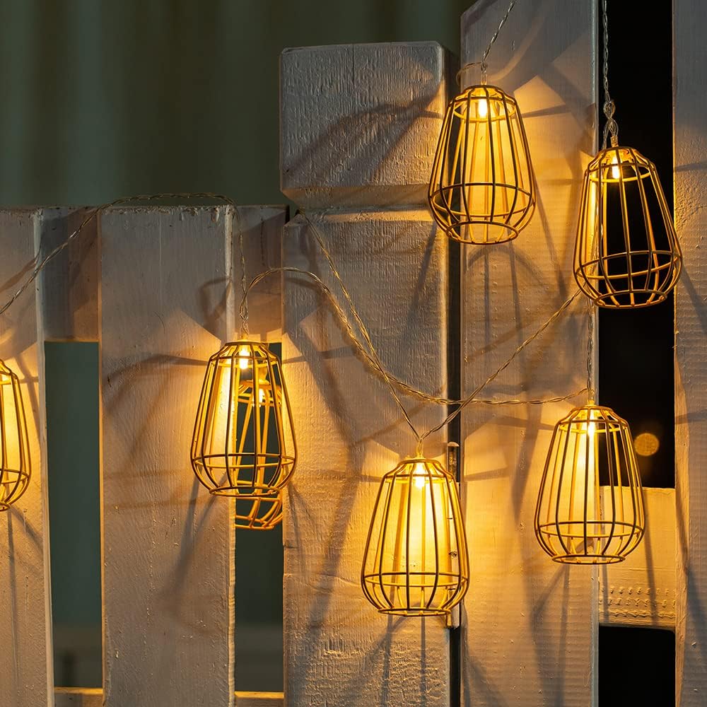 Warm-toned filament bulbs encased in wire cages hang against a white wooden pallet backdrop with sheer curtains, creating a perfect ambiance for an outdoor party.