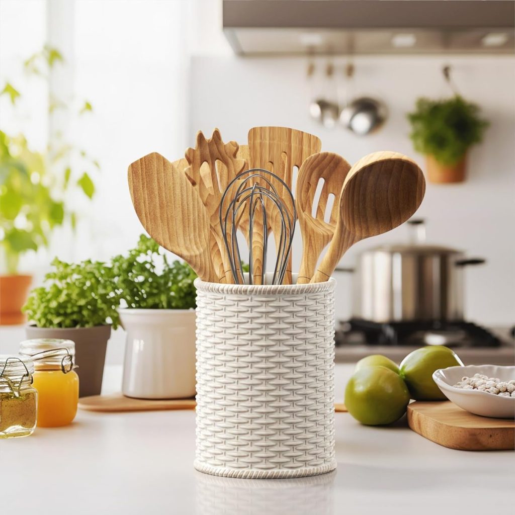 Assorted wooden kitchen utensils in a white woven holder on a countertop with ingredients, spring floral arrangements, and cookware in the background.