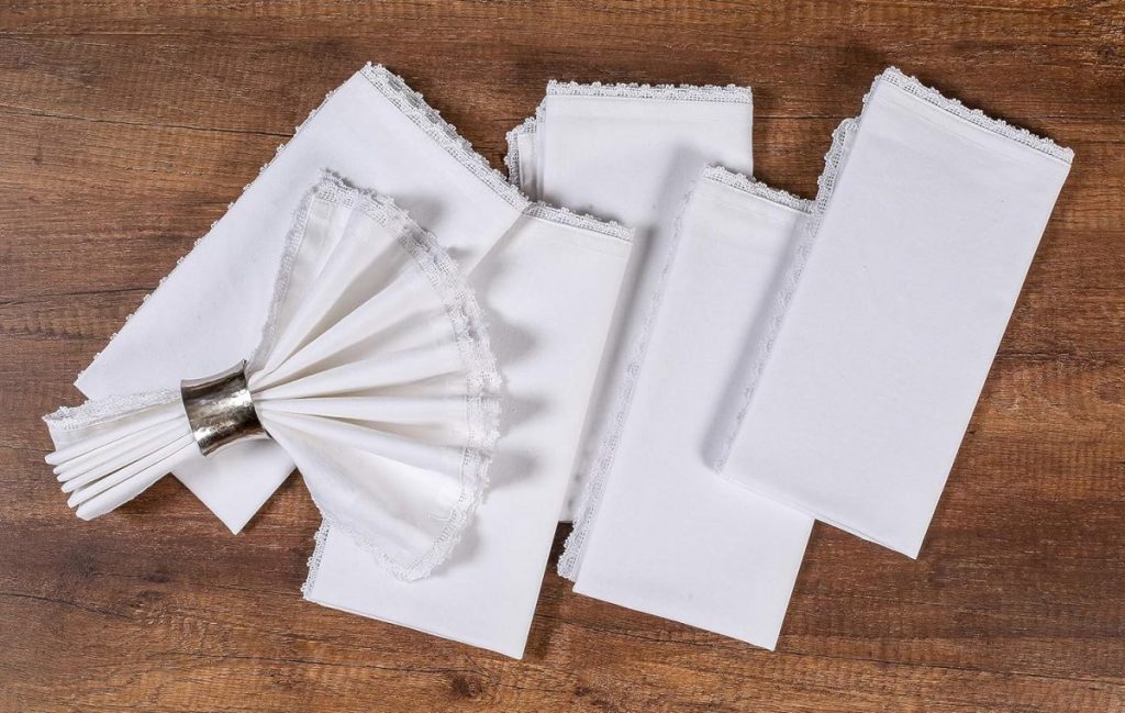 A set of neatly arranged white fabric napkins with decorative borders, partially fanned out on a wooden surface for an outdoor party.