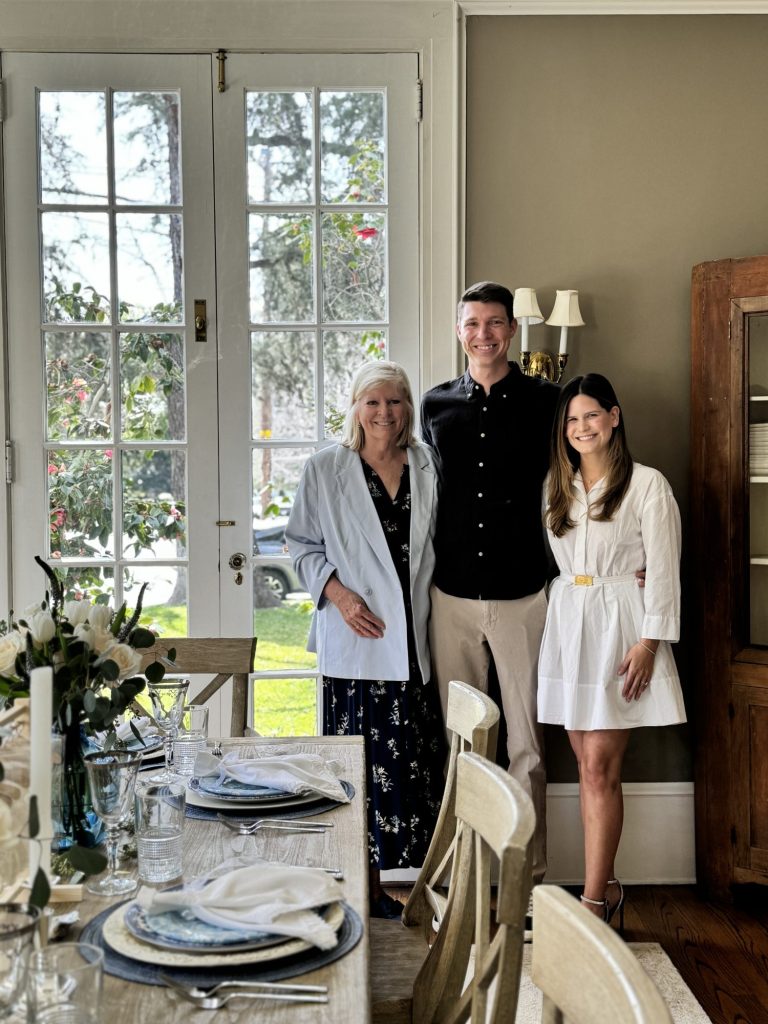 Three people standing by a dining table set for a meal, smiling indoors near french doors.