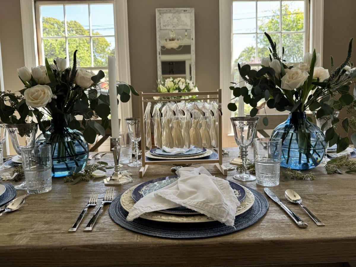 Elegantly set dining table with blue and white tableware, floral centerpieces, and a rack of bridal gown cookies in a bright room.