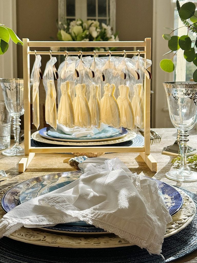 A miniature clothing rack with tiny bridal dress shaped cookes on hangers displayed in front of a window, with a plate and glasses on a dining table in the foreground.