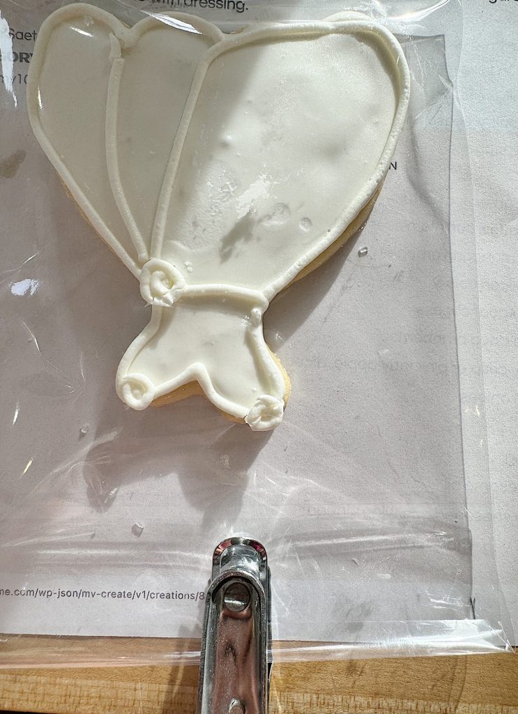 A bridal dress-shaped cookie with white icing next to a metal zipper on a plastic package.