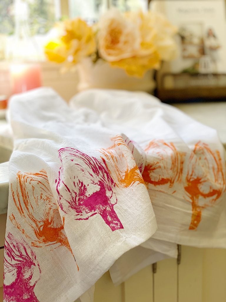 A white linen with vegetable prints laid across a counter in a bright room with yellow flowers in the background.