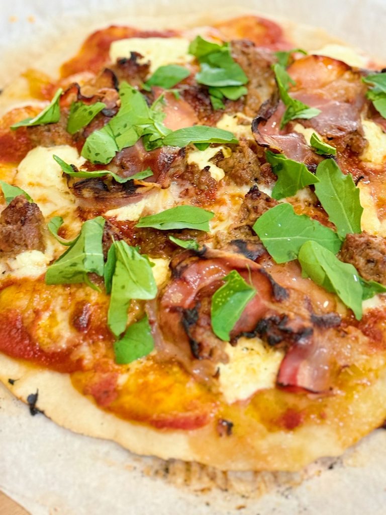Freshly baked pizza with toppings of mozzarella, tomato sauce, sausage, prosciutto, and arugula leaves on a wooden surface.