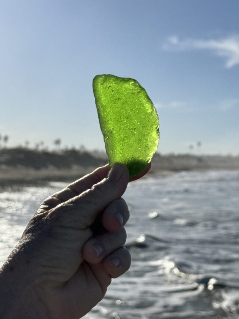 A person holding up a piece of green sea glass with light shining through it against a beach backdrop.