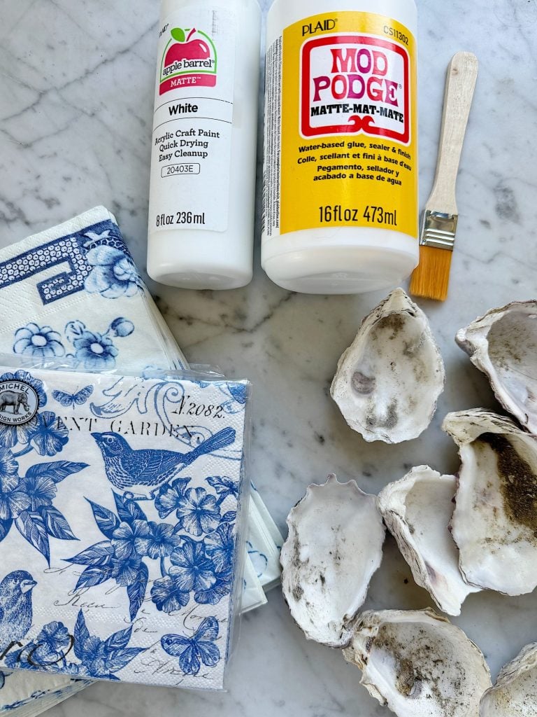 Crafting supplies including white paint, mod podge, a paintbrush, patterned napkins, and oyster shells on a marble surface.