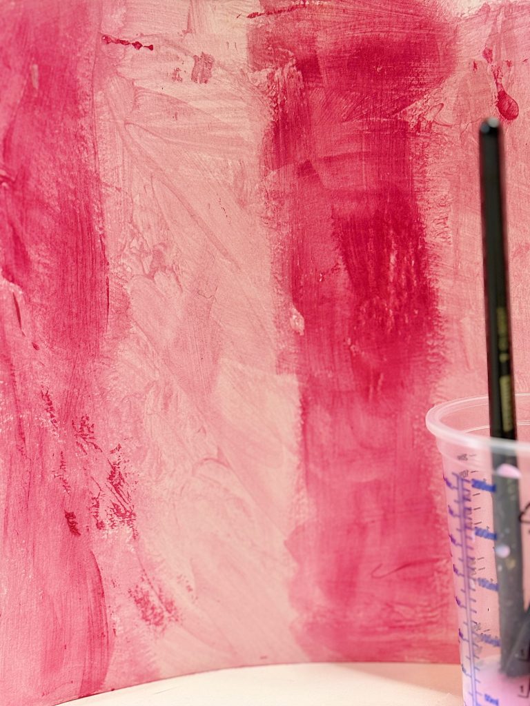 Watercolor paper with abstract pink and dark pink brush strokes next to a paintbrush and a cup with water.
