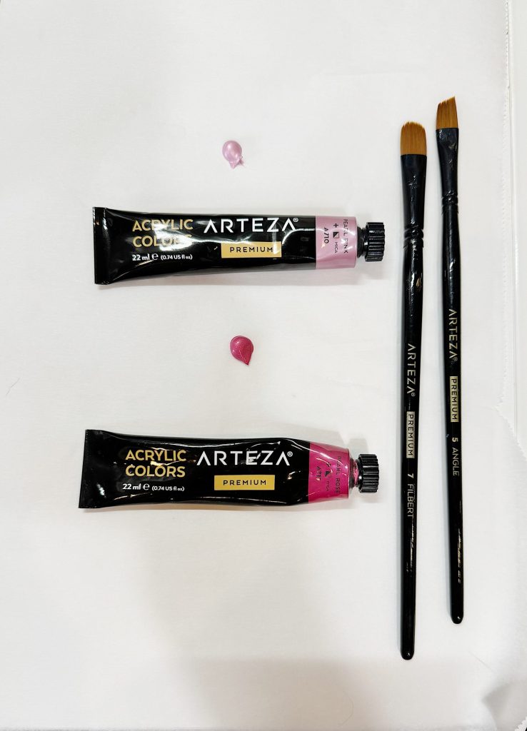 Two tubes of arteza acrylic paint with two paintbrushes on a white surface, with small dabs of pink paint beside each tube.