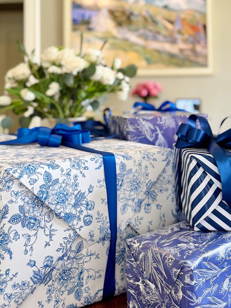 Wrapped gifts with blue and white floral pattern and ribbon on a table, with flowers and a painting in the background.