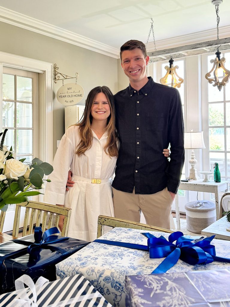 A smiling couple standing in a well-lit room with elegant decor and gifts on the table in front of them.
