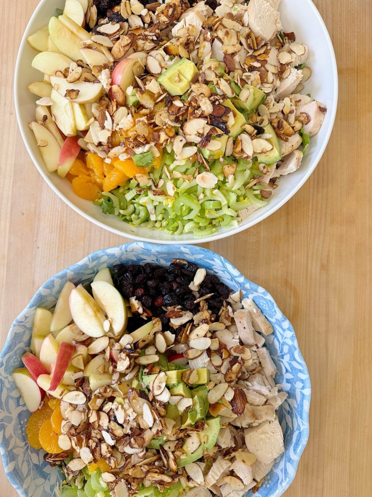 Two bowls of chicken salad with apple slices, mandarin oranges, almonds, and greens on a wooden table.