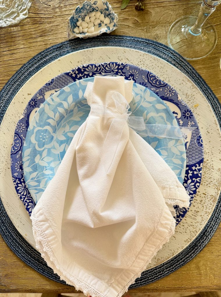 A white napkin folded elegantly like a wedding gown, on a blue patterned plate, set on a wooden table with tableware accents.