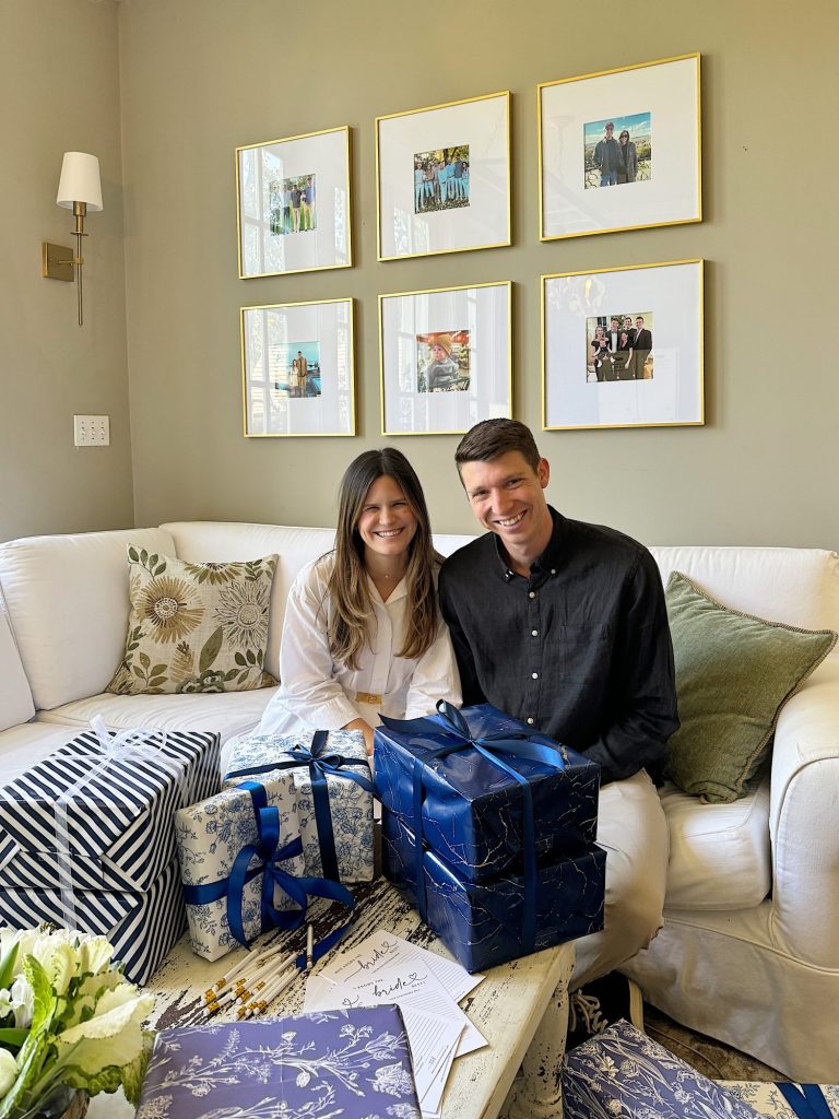 A smiling couple seated on a couch with gifts on their lap and framed pictures on the wall behind them.