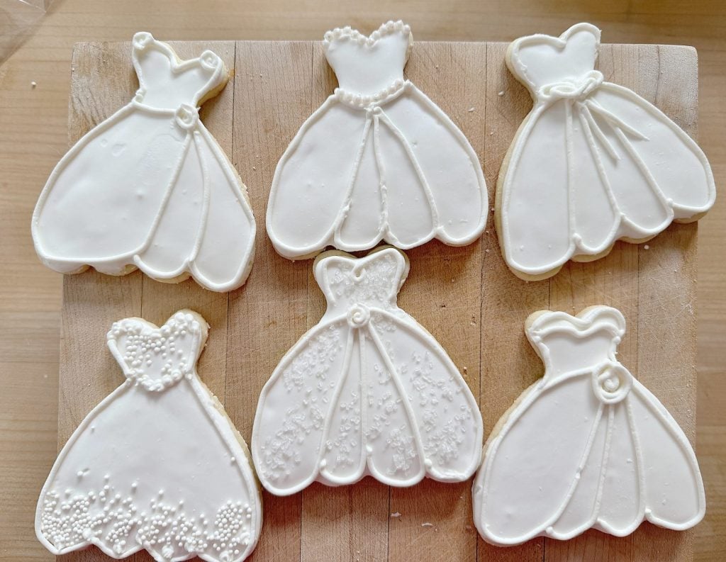 Six wedding dress-shaped cookies with white icing on a wooden board.