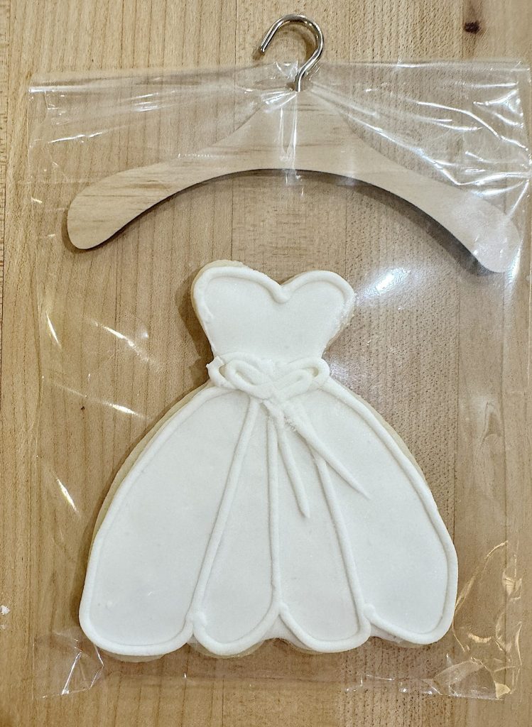 A white dress-shaped cookie attached to a mini wooden hanger, presented in a clear plastic packaging.