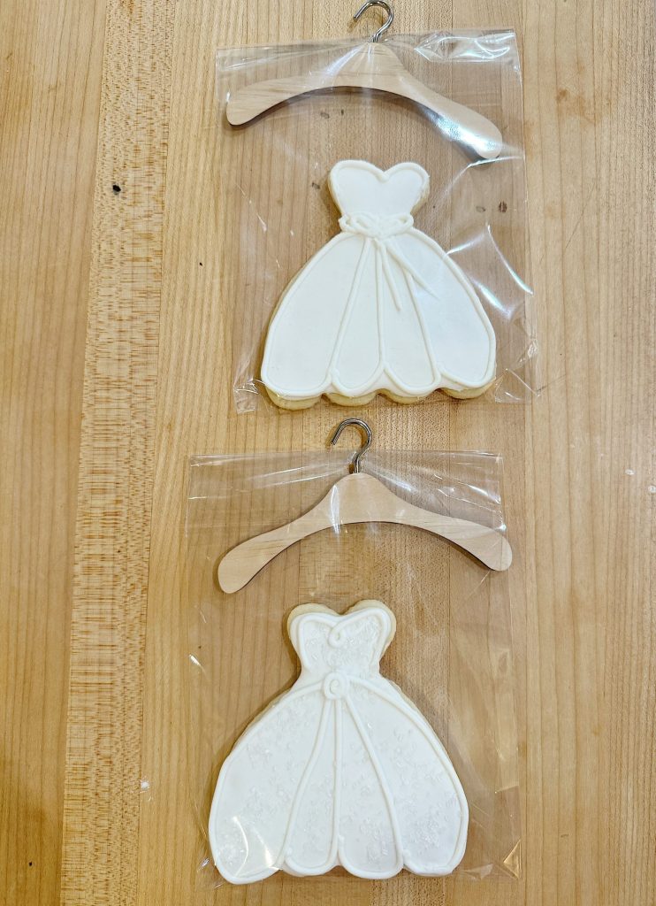 Two white dress-shaped cookies in plastic bags hanging on a wooden surface.