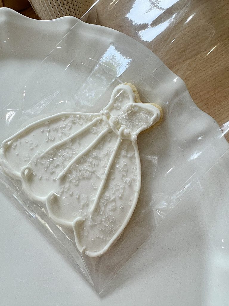 A white frosted cookie shaped like a wedding dress in a plastic bag on a plate.