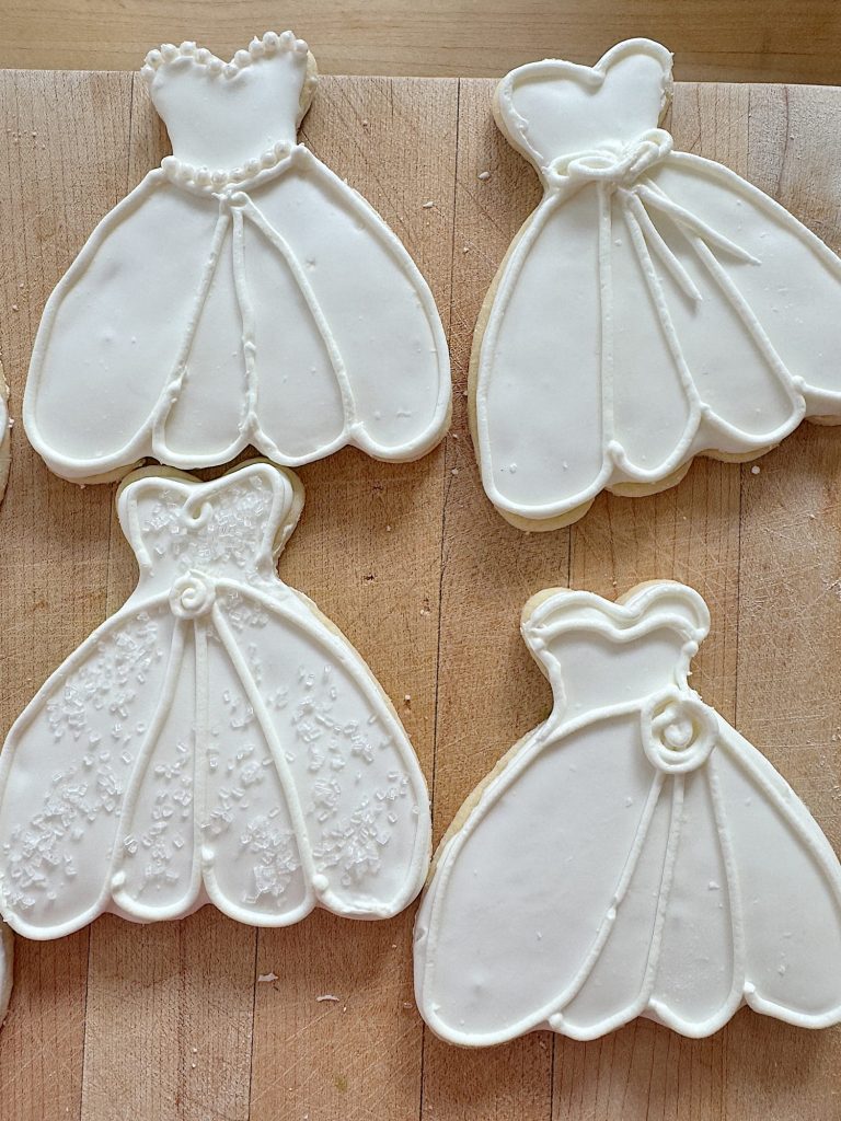 Four wedding dress-shaped cookies with white icing on a wooden board.