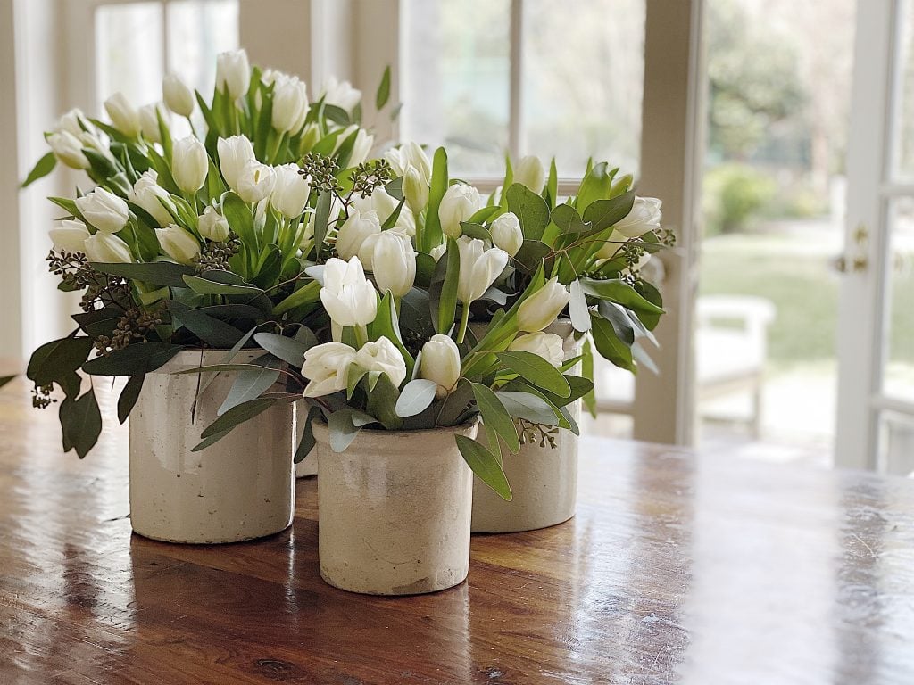 Three rustic vases with white tulips and greenery on a wooden table.