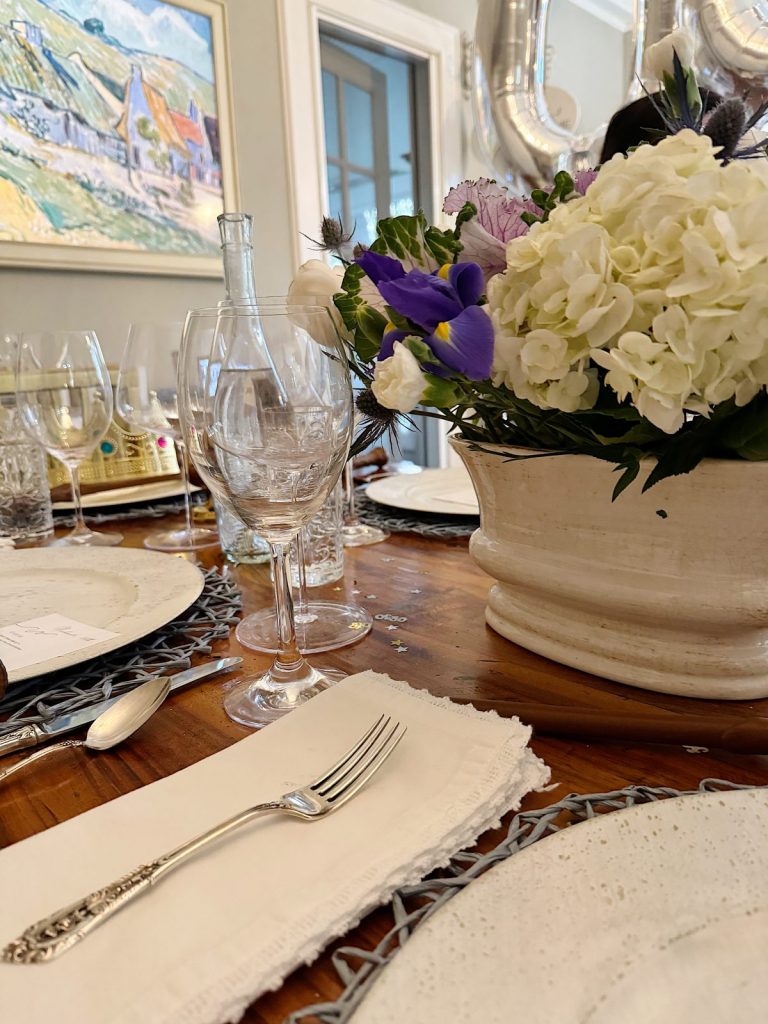 Elegant dining table setting featuring clear wine glasses, silver cutlery, white napkins, and a floral centerpiece, with a painting in the background.