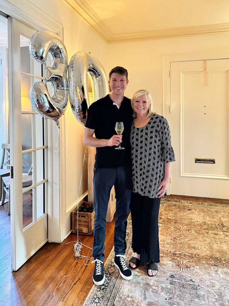 A young man and an older woman smiling, holding a champagne glass next to "30" balloons in a cozy room.