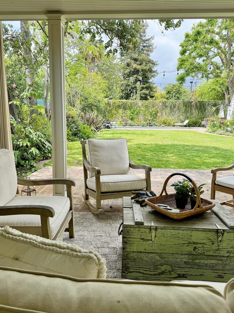 A cozy patio with comfy chairs and a rustic wooden table, overlooking a lush backyard garden visible through wide french doors.