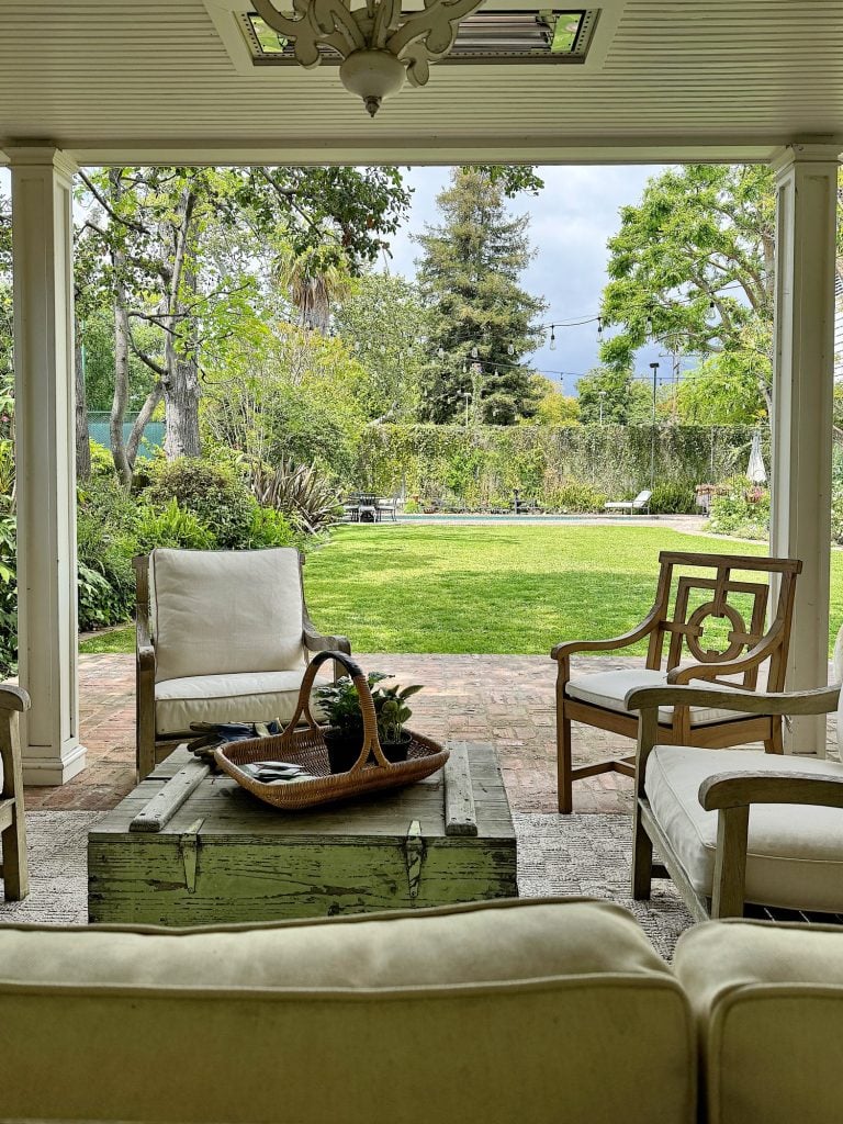 A serene porch with white chairs and a green vintage chest serving as a table, looking out to a lush garden with a mountain backdrop.