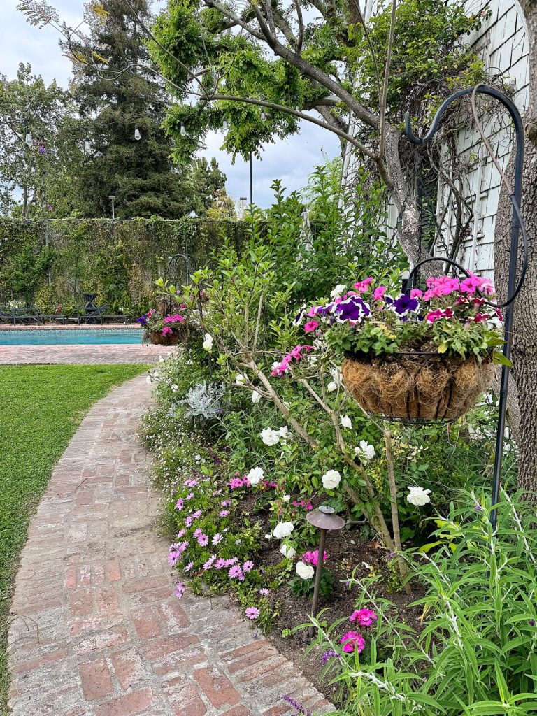 A lush garden pathway with bright pink and white flowers in full bloom, leading towards a pool, complemented by a brick walkway and green trees.