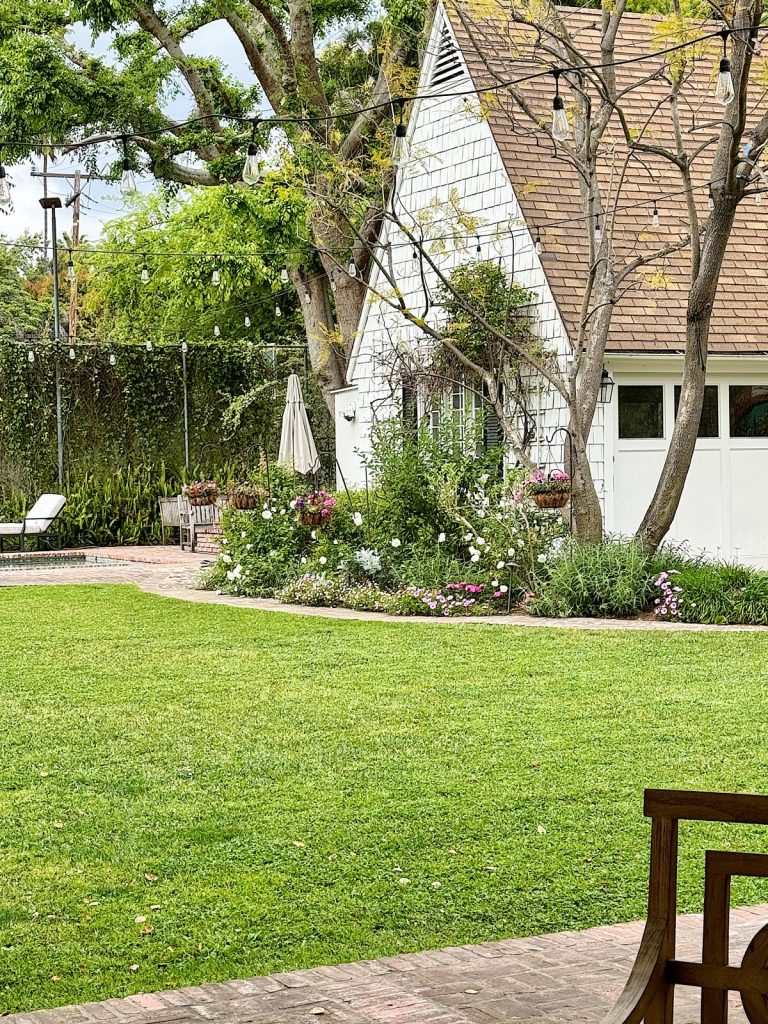 A charming backyard with a well-manicured lawn, colorful flowerbeds, and a white cottage-style house with an a-frame roof.