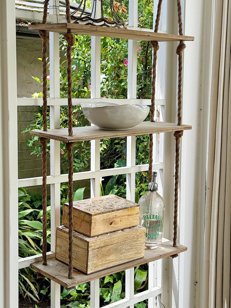 A hanging wooden shelf with ropes, holding a white bowl, old books, and a vintage glass bottle, against a window with a garden view.
