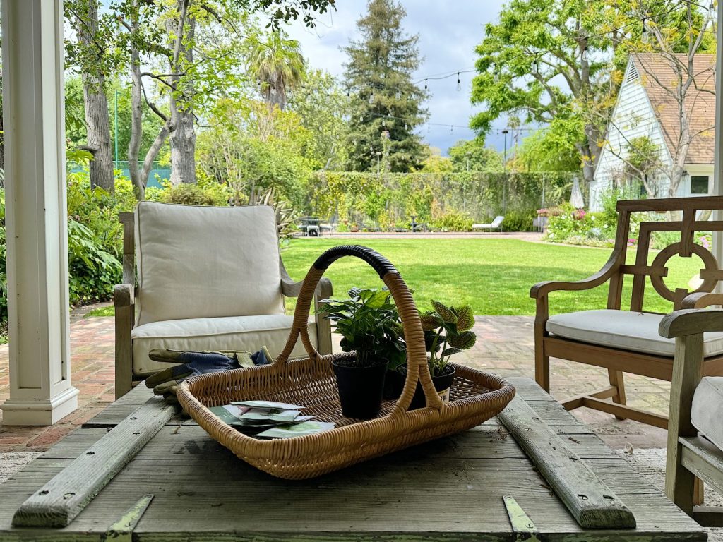 View from a porch with a wicker basket containing plants on a table, overlooking a lush garden and chairs.