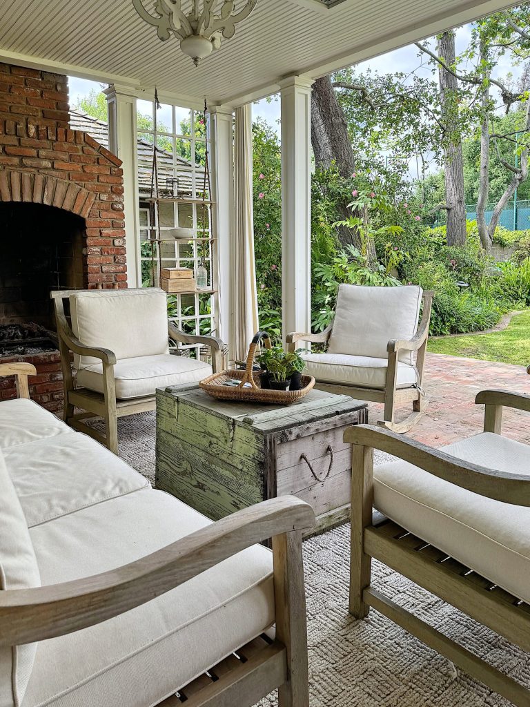 Enclosed patio area with a rustic fireplace, two white cushioned chairs, a wooden table, and a view of a lush garden.